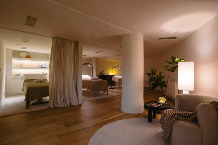 Treatment beds behind linen curtains and a lounge corner with an armchair and floor lamp in a softly lit salon
