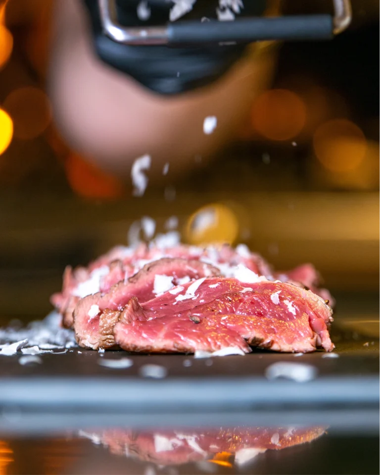 Sliced seared beef on a dark surface in warm, out-of-focus light, with a cook's hand above