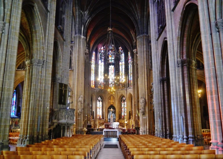 Tall nave with rows of wooden chairs leading to the choir, lit by stained-glass windows and a chandelier