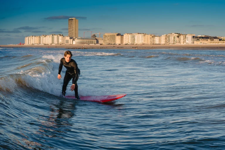 Surfer in a wetsuit riding a small wave, with the Ostend seafront skyline behind