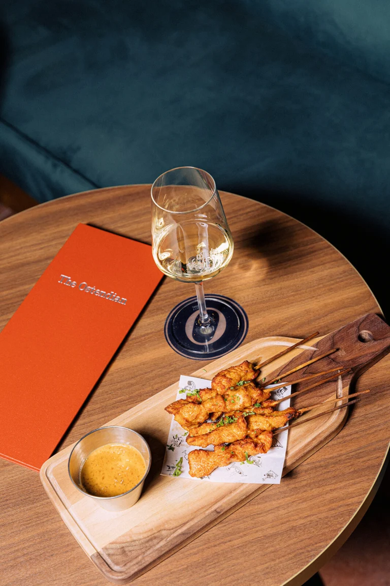 Chicken skewers with dipping sauce on a wooden board next to a glass of white wine and an orange menu on a round table