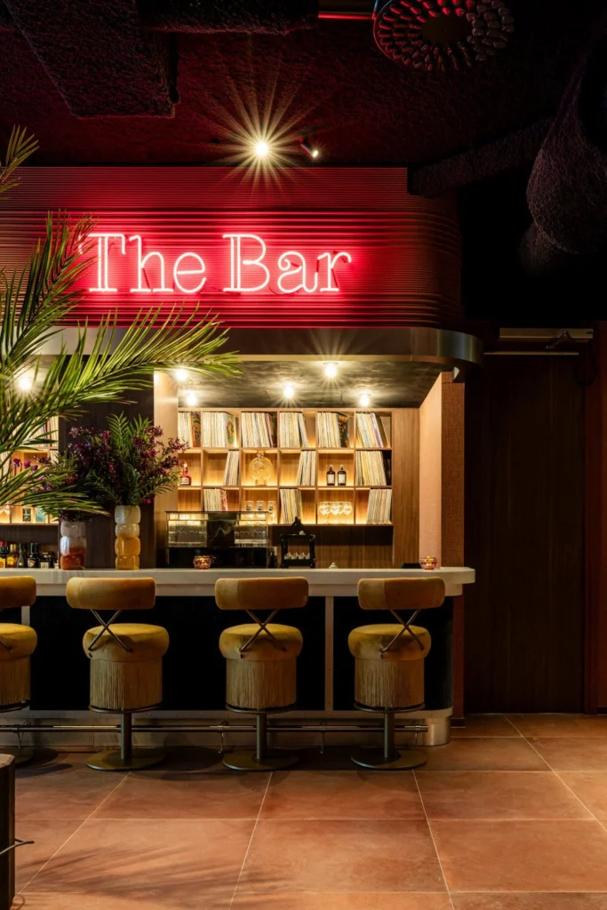 Red neon sign reading The Bar above a counter with fringed bar stools, palm leaves and shelves of vinyl records
