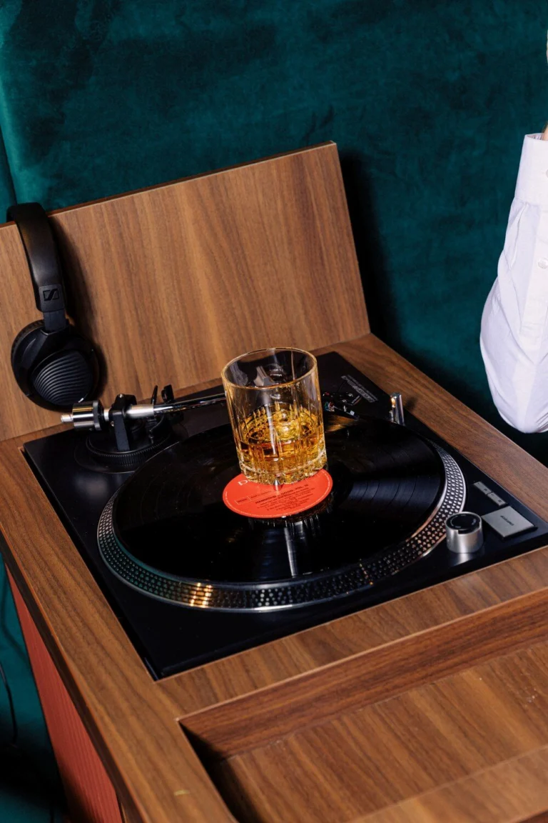 Glass of whisky on ice resting on a record player built into a wooden table, with headphones beside it