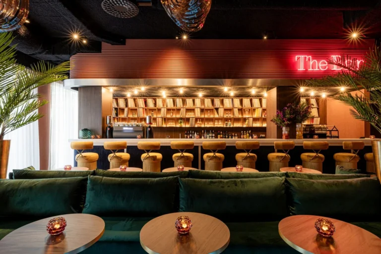 Hotel bar with a red neon ‘The Bar’ sign, rows of stools and green velvet banquettes with candlelit tables
