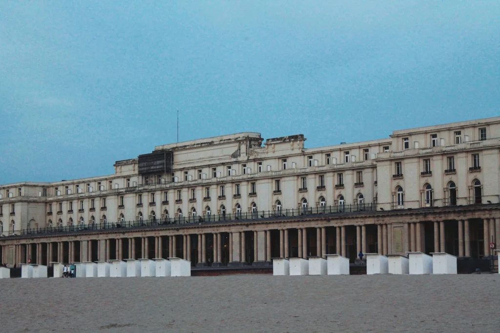 Long cream facade of the Thermae Palace and Royal Galleries seen from the beach, with white beach cabins in front