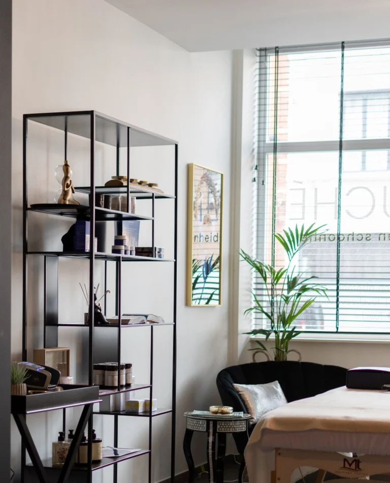 Bright treatment room with a massage table, open metal shelving with products and plants by the window