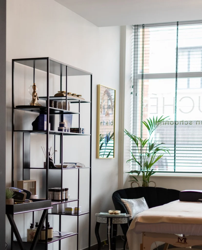 Bright treatment room with a massage table, open metal shelving with products and plants by the window