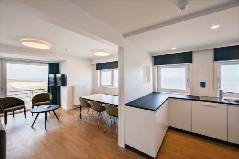 Apartment with a white kitchenette, dining table, two armchairs and windows looking out towards the beach