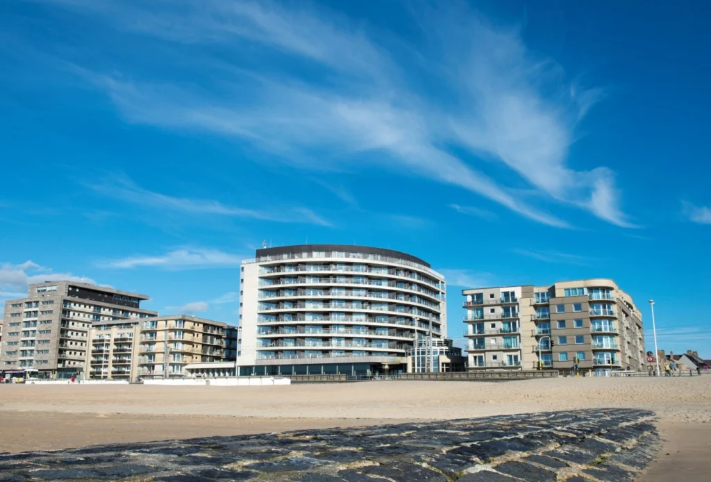 Curved white Vayamundo building on the seafront between apartment blocks, seen from the beach under a blue sky