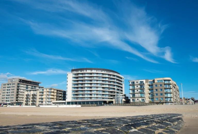 Curved white Vayamundo building on the seafront between apartment blocks, seen from the beach under a blue sky