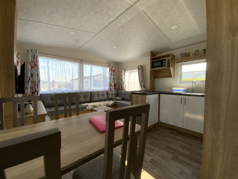 Mobile home interior with a dining table, corner sofa and fitted kitchen beneath wide windows