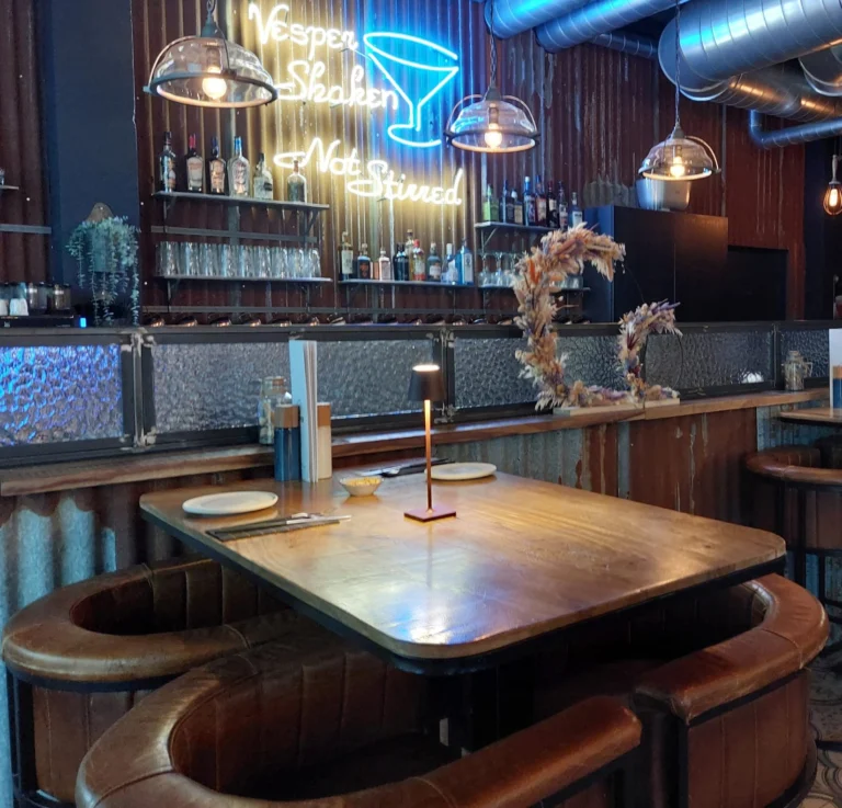 Leather booth and wooden table beneath a blue neon sign reading 'Vesper — Shaken, not stirred' with a cocktail glass, in front of the bar