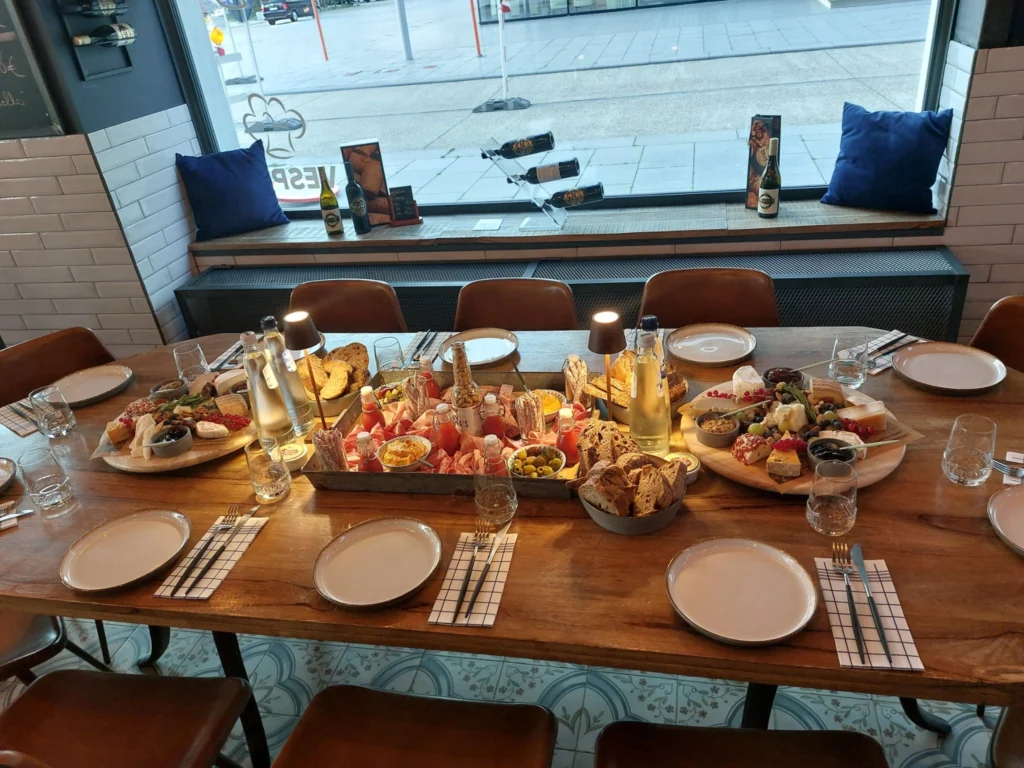 A wooden table laid for a group, covered with sharing boards of charcuterie, cheese, seafood and bread, by a window onto the quay