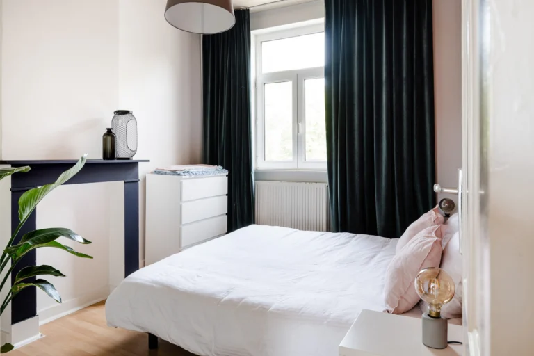 Light bedroom with a white double bed, dark green velvet curtains and a painted fireplace surround