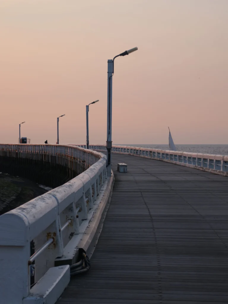 The curving wooden boardwalk and white railings of the West Pier at dusk, with a sailing boat offshore
